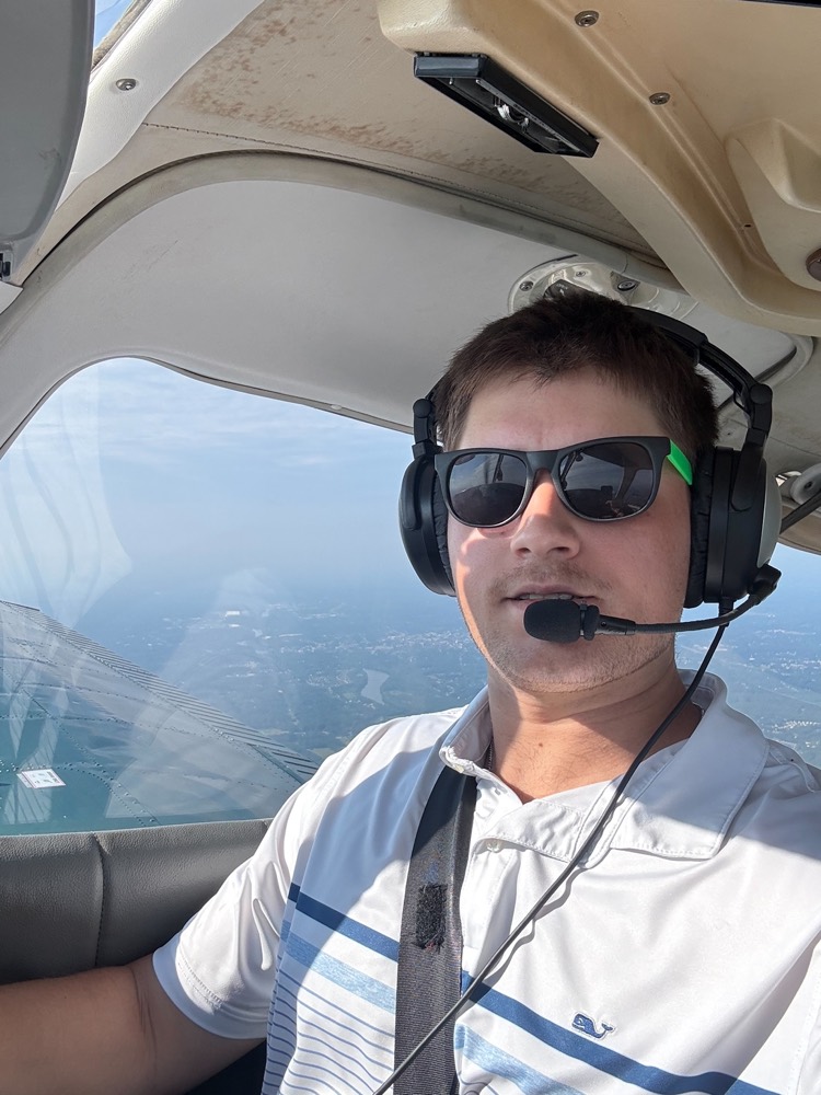 Adam Clark, CFI-I, smiling in the cockpit wearing a headset and sunglasses during a flight
