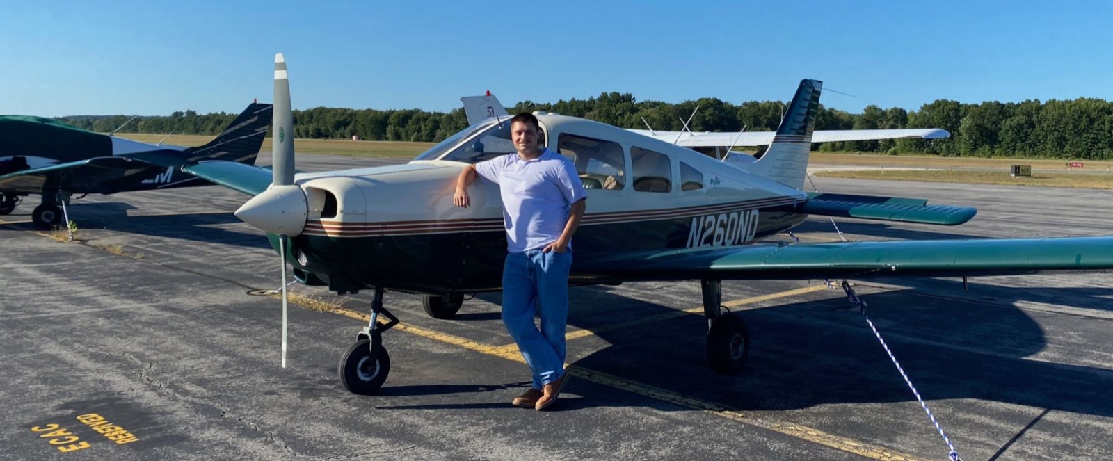 Adam Clark leaning against the nose of a white and green Piper airplane, tail number N260ND, on the ramp under a clear blue sky